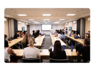 Sala de reunião com fileiras de mesas e participantes sentados diante de uma tela de projeção